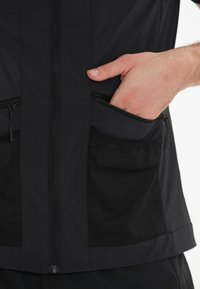 Svart jacka i ett mjukt tyg med nätinslag, med dragkedjefickor och en mittdragkedja. Närbild av fickor och hand.