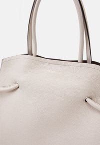 Beige läder toteväska med en texturerad yta, featuring två handtag upptill och en subtil guldlogotyp på framsidan. Enkelt och funktionellt design.