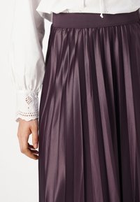 Gonna midi plissettata di un viola scuro con una ampia vita elastica, realizzata in un tessuto lucido e liscio. Blusa bianca ricamata con polsini in pizzo è parzialmente visibile.