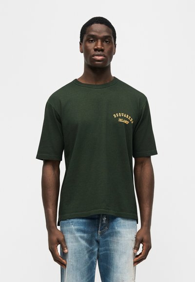 Άνδρας με κοντά μαλλιά που φοράει σκούρα πράσινη μπλούζα DSQUARED2 ISLAND® και ανοιχτόχρωμο ξεθωριασμένο τζιν, στέκεται μπροστά σε απλό φόντο.
