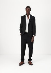 Black suit with notched lapels, white shirt, and black trousers. The outfit features a relaxed fit and brown leather shoes with a smooth texture.
