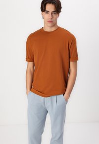 T-shirt en coton marron avec des manches roulées, associé à un pantalon plissé bleu clair. Design simple, col rond et texture douce.