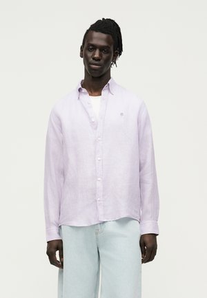 Άνδρας με σκούρο δέρμα και κοτσιδάκια, φοράει ένα ανοιχτό μωβ πουκάμισο πάνω από λευκό T-shirt και ανοιχτό μπλε τζιν, στέκεται μπροστά σε απλό φόντο.