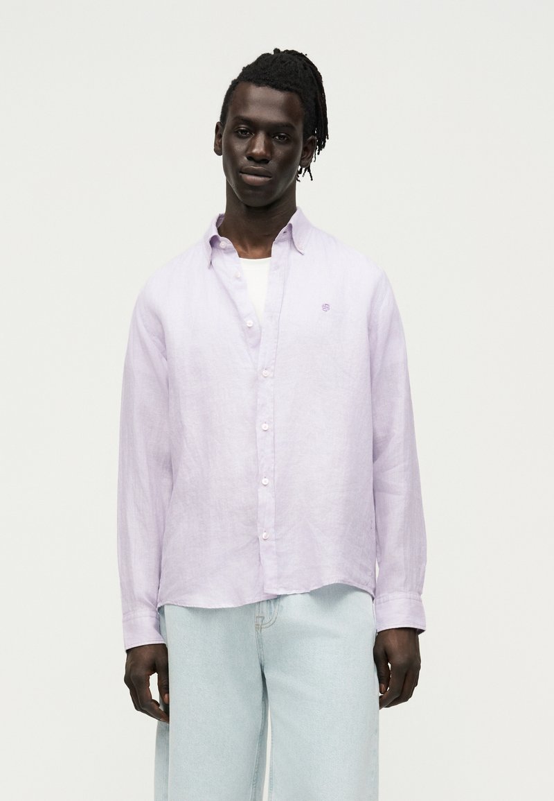 Άνδρας με σκούρο δέρμα και κοτσιδάκια, φοράει ένα ανοιχτό μωβ πουκάμισο πάνω από λευκό T-shirt και ανοιχτό μπλε τζιν, στέκεται μπροστά σε απλό φόντο.