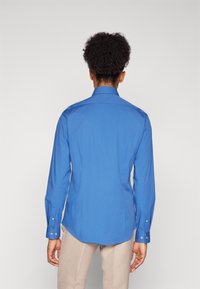 Camicia azzurra a maniche lunghe con colletto classico, vestibilità su misura e polsini con bottone. Tessuto liscio con un leggero luccichio; vista posteriore che mostra l'orlo curvato.