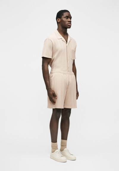 Homme debout portant une chemise beige à manches courtes avec col et un short assorti, accompagnés de chaussettes beiges et de baskets blanches, sur fond blanc.