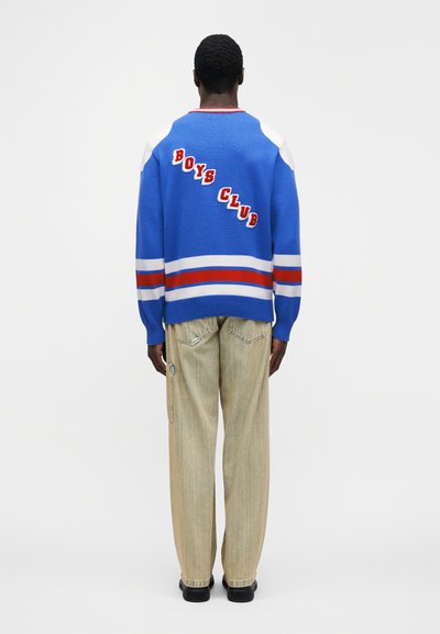 Μπλε πλεκτό πουλόβε ρ με το κείμενο "BOYS CLUB" στην πλάτη, με κόκκινες και λευκές ρίγες στα μανίκια. Συνδυάζεται με ανοιχτό μπεζ τζιν παντελόνι φαρδιάς γραμμής.