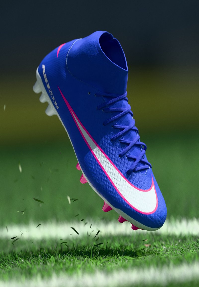 Mėlynas futbolo batelis su aukštu apykakliu, baltas ir rožinis „Swoosh“ logotipas, tekstūruotas viršus ir rožinės smeigės, pakeltas virš žolės.