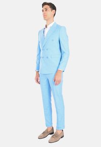 Giacca blu chiaro doppiopetto con finitura texturizzata, abbinata a una camicia bianca e a scarpe in suede beige, con vestibilità sartoriale.