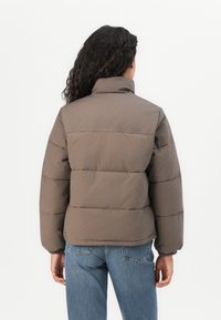 Giacca imbottita marrone con colletto alto, polsini elasticizzati e vestibilità ampia. Tessuto liscio, senza motivi visibili. Indossata sopra dei jeans blu.