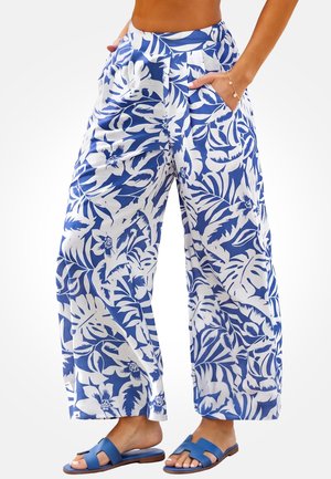 Femme portant un pantalon blanc à jambes larges avec imprimé floral tropical bleu et des sandales plates ouvertes bleues, mains dans les poches.