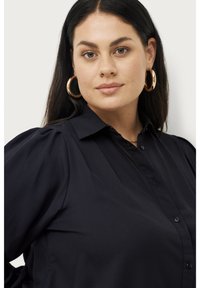 Navyblauw overhemd met knopen en pofmouwen, gemaakt van een gladde stof. Model draagt grote gouden oorbellen. Zachte textuur, minimalistisch ontwerp.