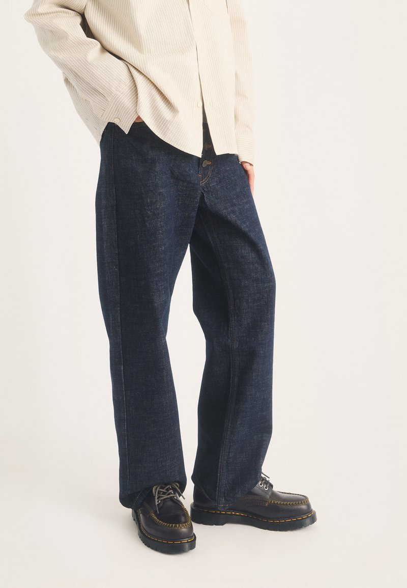 Personne portant un jean bleu foncé à jambes larges, des chaussures noires à lacets avec des coutures jaunes, et une chemise beige clair à manches longues, les mains dans les poches.