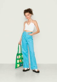 Top halter branco, jeans de cintura alta azuis e sandálias de plataforma pretas. Bolsa tote verde com padrões de flores coloridas. Fundo simples.