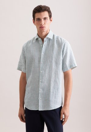 Jeune homme portant une chemise à manches courtes boutonnée, rayée verticale verte et blanche, avec un pantalon foncé, debout devant un fond uni.