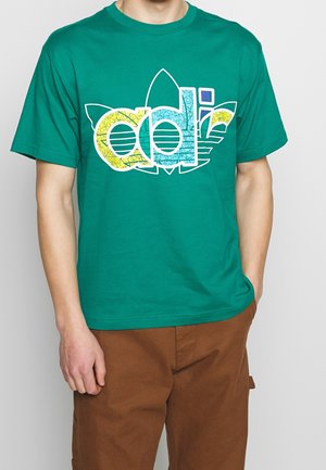 Homme portant un t-shirt vert avec un logo Adidas multicolore et un pantalon marron, debout avec une main dans la poche devant un fond uni.