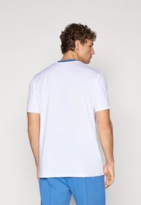 Hvid atletisk t-shirt med blå accent ved halsen. Korte ærmer og glat, letvægtigt stof. Enkel design, set bagfra.