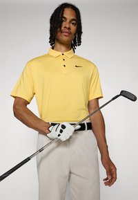 Κίτρινο κοντομάνικο μπλουζάκι polo με τρία μαύρα κουμπιά και μικρό μαύρο λογότυπο, συνδυασμένο με ανοιχτόχρωμο παντελόνι και κρατώντας ένα μπίλια γκολφ.