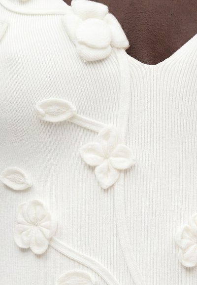 Άσπρο ριμπ ύφασμα με ανάγλυφα ανθικά εφαρμογές σε ίδιο λευκό χρώμα κατά μήκος  μιας καμπυλωτής ραφής κοντά σε σκούρο δέρμα στο ντεκολτέ.