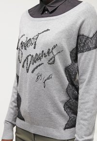 Pull en gris avec des panneaux en dentelle noire, texte en sequins disant "Sweet Dreams", et poignets côtelés. Présente un décolleté décontracté et tombant sur une épaule.