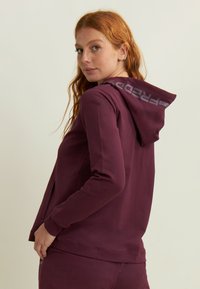 Donna con lunghi capelli rossi, che indossa un hoodie bordeaux e pantaloni abbinati, che guarda oltre la spalla su uno sfondo neutro.