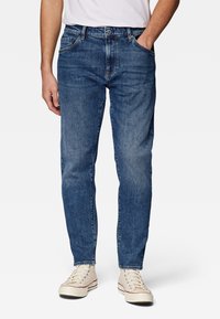 Mies, joka käyttää sinisiä farkkuja ja vaaleita kangaskengät, toinen käsi taskussa, seisoo yksinkertaista valkoista taustaa vasten.