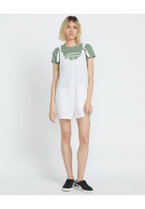 Hvid linned overalls kjole med justerbare stropper, frontlommer, båret over en grøn t-shirt med et hvidt grafik. Sorte sneakers fuldender looket.