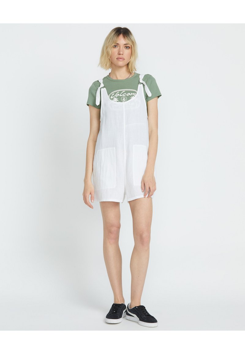 Abito intero bianco in lino con spalline regolabili, tasche frontali, indossato sopra una maglietta verde con un motivo bianco. Scarpe da ginnastica nere completano il look.
