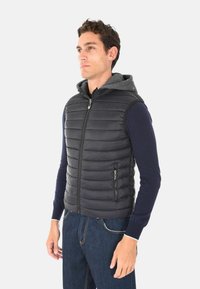 Uomo che indossa un gilet senza maniche in tessuto trapuntato nero con cappuccio grigio sopra una maglietta a maniche lunghe blu navy e jeans blu, in piedi contro uno sfondo bianco.