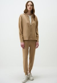Beige gebreide hoodie met een rits en een trekkoord, gecombineerd met bijpassende taps toelopende broeken, beide gemaakt van zachte stof. Witte sneakers maken de look compleet.