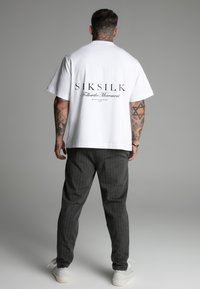 Fehér rövid ujjú póló, hátulján fekete felirattal: "SIKSILK Follow the Movement". Szürke csíkos nadrággal és fehér cipővel párosítva.
