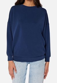 Felpa blu navy, vestibilità comoda, scollo tondo, maniche lunghe con polsini a coste, texture morbida, indossata con jeans azzurri.