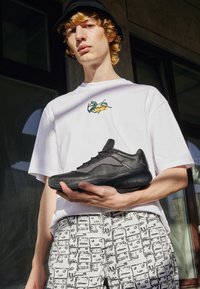 Scarpa sportiva nera con superficie texturizzata e dettagli in rete, tenuta da una persona che indossa una camicia bianca con ricami floreali e pantaloni chiari a fantasia.