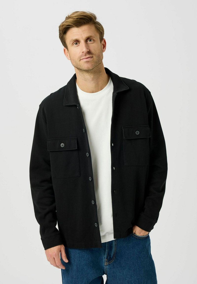 Chemise noire boutonnée avec deux poches avant, col à revers et tissu doux, associée à un t-shirt blanc et un jean bleu.