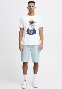 Hvit bomulls-T-skjorte med et fargerikt grafisk trykk av en fisk med kapteinslue, sammen med lyseblå denimshorts og hvite joggesko.