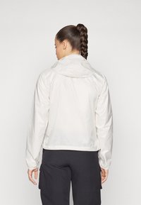 Giacca leggera bianca con cappuccio, caratterizzata da una texture liscia e polsini elasticizzati, indossata sopra pantaloni neri, vista da dietro.