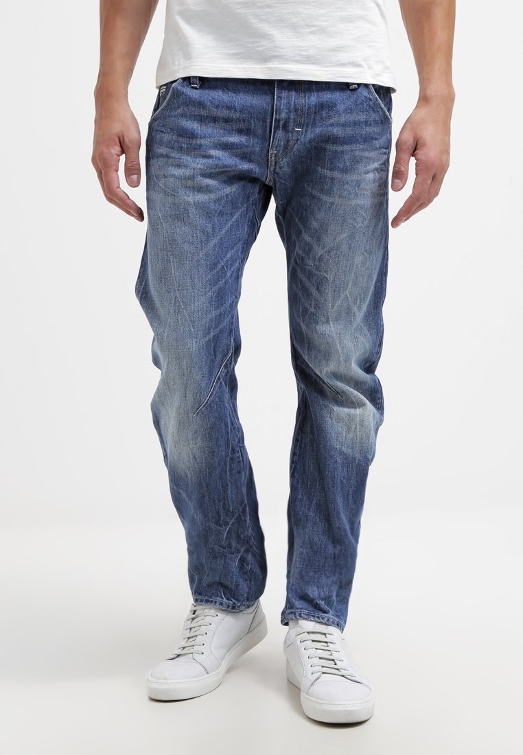 Homme portant un jean bleu délavé et des baskets blanches, debout contre un fond blanc uni, les mains détendues le long du corps.