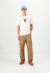 White t-shirt with black and red logo, khaki pants, and orange shoes. Light blue cap completes the outfit; standing against a plain background.