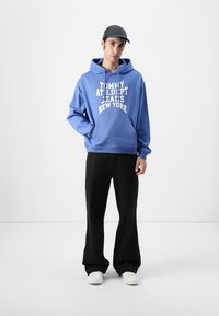 Blue hoodie with white text “TOMMY ATH. DEPT. JEANS NEW YORK,” front pocket; paired with black pants and white sneakers.