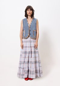 Gestreifter blauer Weste mit beigem Saum und drei Knöpfen, kombiniert mit einem gestuften karierten Maxikleid in Hellrosa, Grau und dunklen blauen Akzenten. Rote Schuhe.