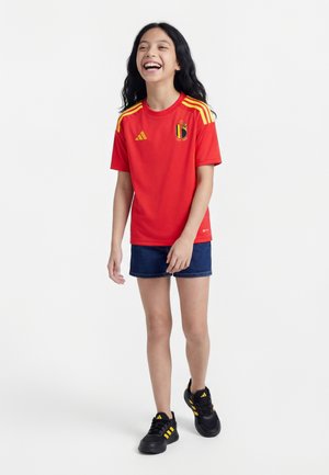 Młoda dziewczyna śmieje się, ubrana w czerwoną belgijską koszulkę piłkarską z żółtymi paskami, dżinsowe szorty i czarne buty sportowe Adidas z żółtymi paskami.