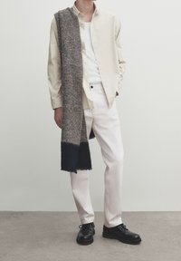 Beżowa koszula polo, biała koszulka na ramiączkach, jasne spodnie i czarne buty. Szary wełniany szalik z ciemnoniebieskimi końcami zwisa z jednego ramienia.