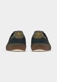 Μαύρα και ελιά πράσινα παπούτσια με υφασμένες λαστιχένιες σόλες, με χρυσό λογότυπο Adidas στη φτέρνα και πλευρά. Κανονικό κλείσιμο με κορδόνια, σχεδίαση χαμηλού κορμού.