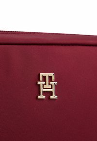Borsa in tessuto borgogna con logo metallico dorato lucido con le iniziali doppie "T" e "H". Texture liscia con dettagli di cucitura precisi.