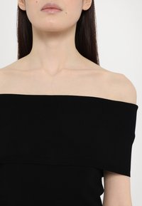 Haut noir en maille côtelée à épaules dénudées, avec un décolleté droit et une texture lisse, conçu pour une silhouette ajustée.