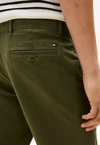 Chino in cotone verde oliva con una tasca posteriore dotata di patch con logo. Il tessuto ha una consistenza liscia e una chiusura a bottoni sulla tasca.