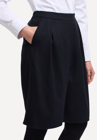 Culottes blu navy in misto lana con pieghe, vita elasticizzata e tasche laterali, abbinati a una camicia bianca. Tessuto liscio, design al ginocchio.