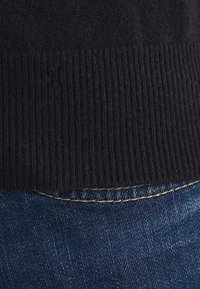 Svart ribbad stickad tröja med fåll ovanför mörka denimjeans, med subtil sömnad längs midjan och en mjuk textur.