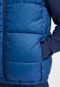 Zils pūkaina veste ar izšūtu dizainu, ar rāvējslēdzēju, sānu kabatām un kontrastējošu teksturētu audumu uz rokām.