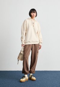 Sweatshirt bege com capuz, calças cargo castanhas com bolsos e ténis bege com atacadores pretos. A carregar uma bolsa bege clara.